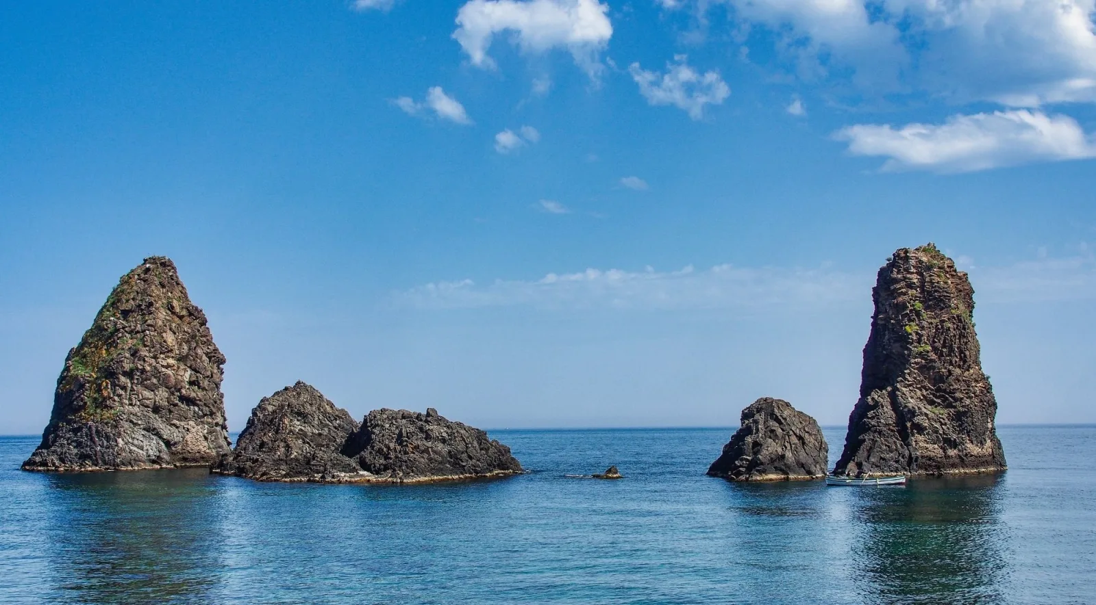 A small fishing boat dwarfed by the tall basalt sea stacks of the Riviera dei Ciclopi