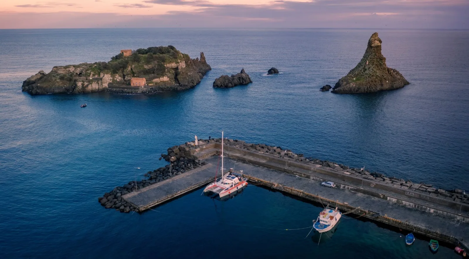 Aerial view at dusk of Isola Lachea, the Faraglioni and a catamaran moored at the Aci Trezza pier