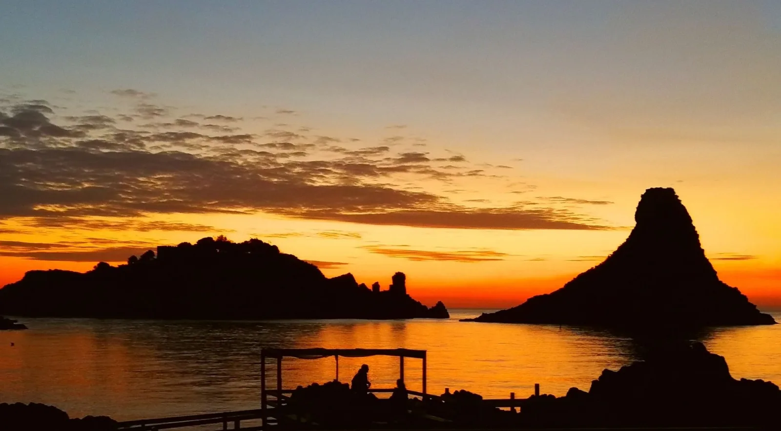 Silhouette of the Faraglioni dei Ciclopi against an orange dawn sky over Aci Trezza