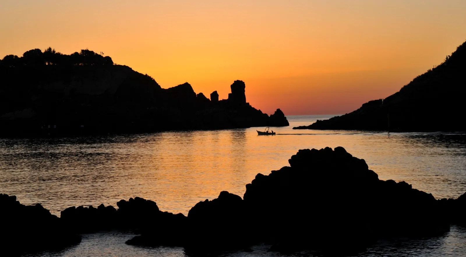 A small boat crossing the calm bay at Aci Trezza at first light, with the rocks silhouetted